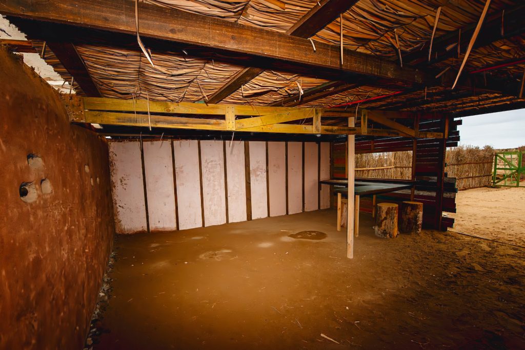 Interior del refugio techado, con suelo de arena y vigas de madera